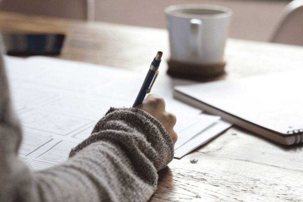 Person writing on paperwork at a wooden desk with a notebook and coffee mug nearby